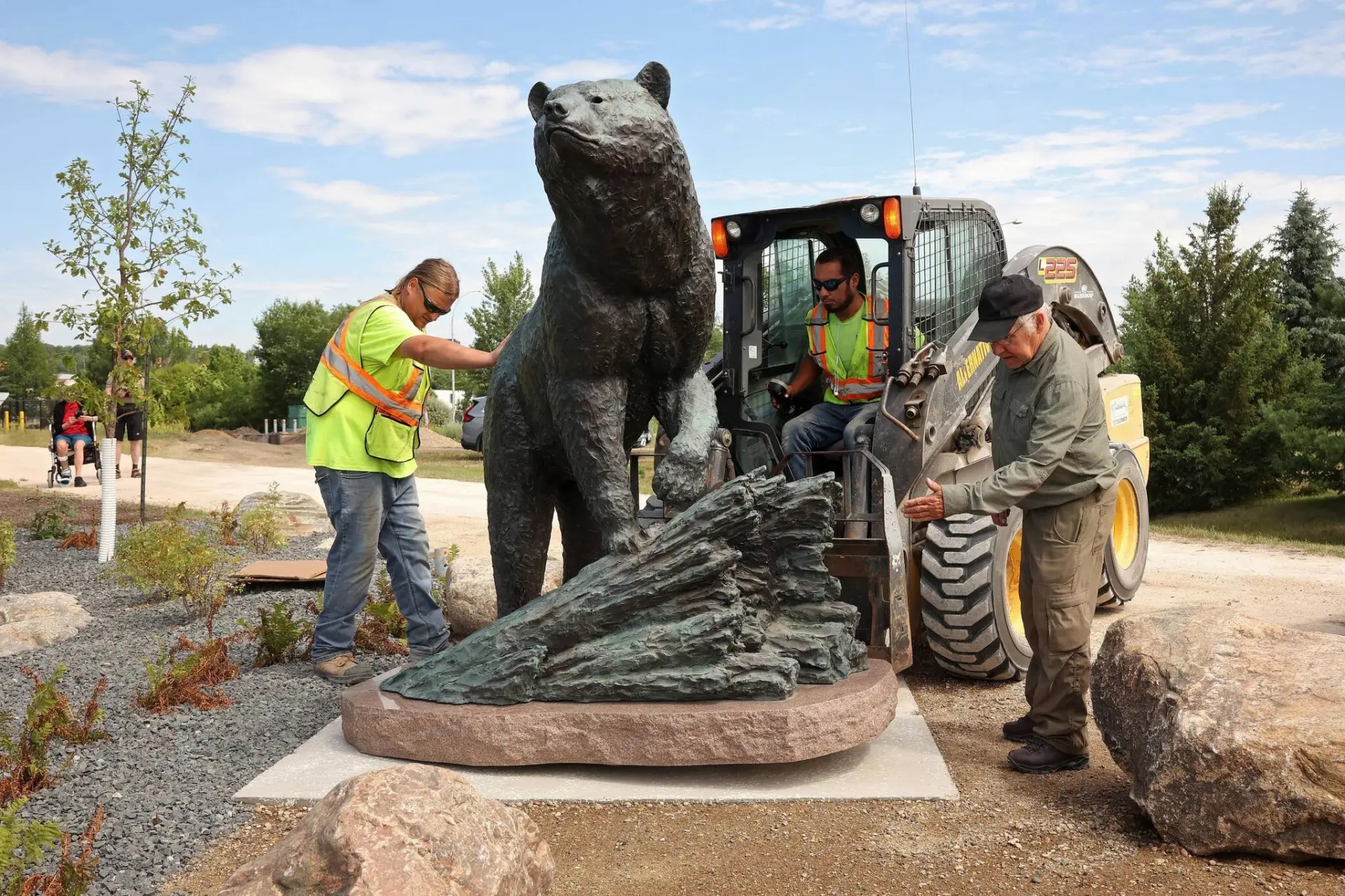 Three men, including Peter Sawatzky and one driving a skid-steer install a large bronze bear sculpture in a park