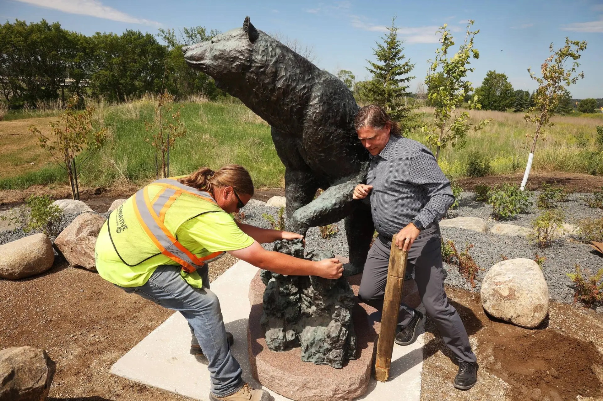 Two men install a large bronze sculpture of a black bear in a park