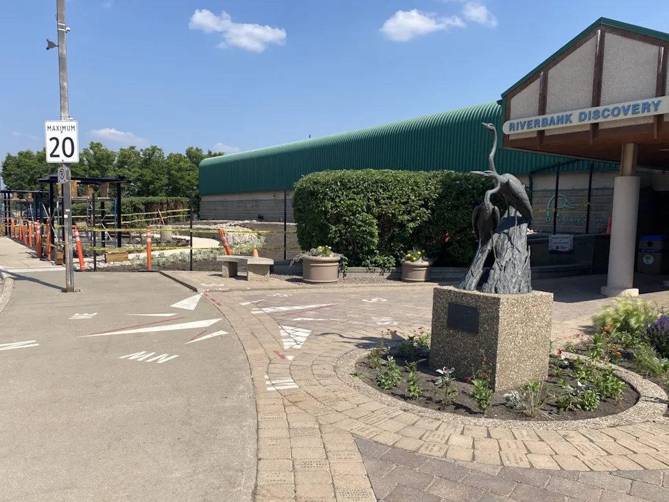 Photo of a building with the sign "Riverbank Discovery" and a bronze sculpture of two herons out front, and flower beds under construction in the background